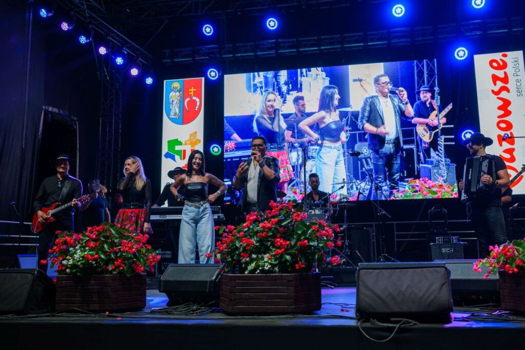 Scena z zespołem muzycznym i dużym ekranem w tle. Kolorowe reflektory i czerwone kwiaty dekorują scenę. Tłem jest nocne niebo.