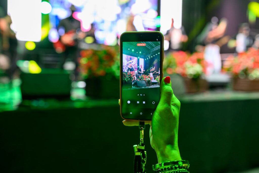 Telefon z czerwonym nagrywaniem kadruje koncert na scenie z zielonymi światłami. W tle rozmazane postacie i kolorowe dekoracje. Ręka trzymająca telefon ma pomalowane paznokcie.