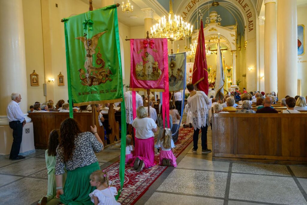 Procesja w kościele z kolorowymi sztandarami, otoczona ludźmi w jasnych ubraniach. Wnętrze jest jasne, z dużymi żyrandolami. Na marmurowej posadzce widać czerwony dywan.