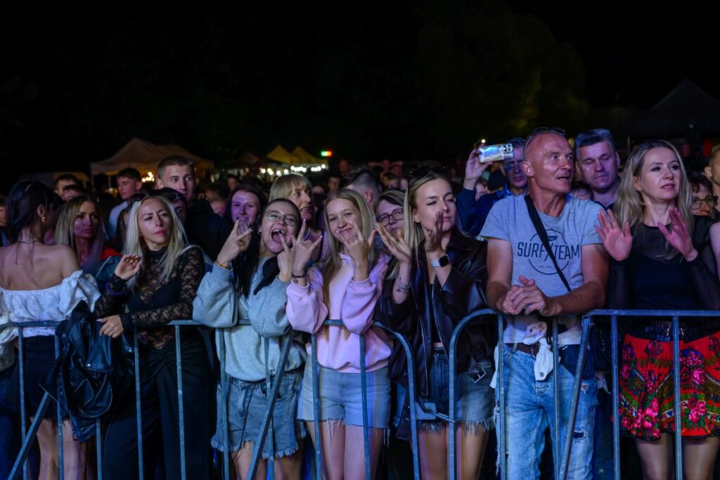 Grupa ludzi stoi za barierką na koncercie nocą. Uśmiechnięte twarze i kolorowe, codzienne ubrania. W tle ciemne niebo i namioty.