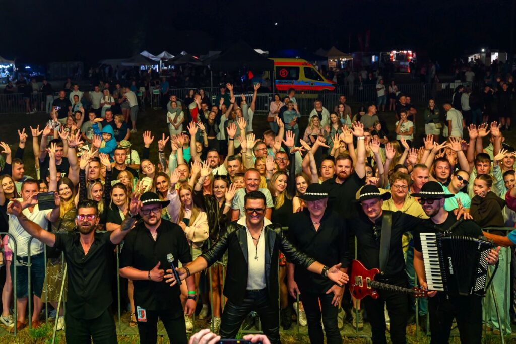 Na zdjęciu widzimy tłum ludzi pod sceną, a na pierwszym planie mężczyzn z instrumentami. Wszyscy są uśmiechnięci, podnoszą ręce. Tło jest ciemne, z widoczną kolorową karetką i stoiskami w oddali.
