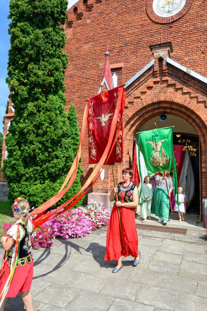 Grupa ludzi w tradycyjnych strojach niesie kolorowe sztandary przed ceglaną ścianą kościoła, w tle kwiaty i drzewo.