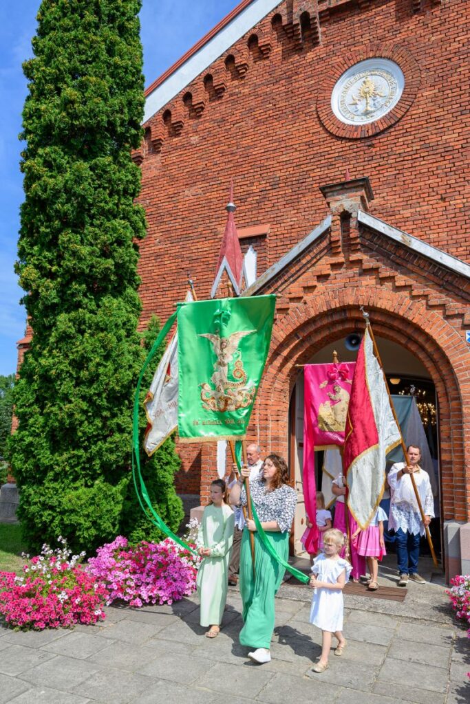Grupa osób niesie kolorowe sztandary przed ceglaną fasadą kościoła. Otoczenie zdobią różowe kwiaty i zielony krzew. Tło jest jasne i słoneczne.