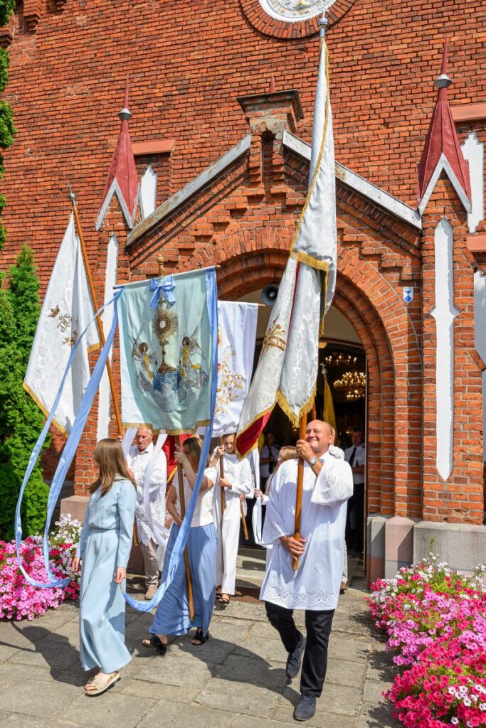Procesja religijna przed ceglanym kościołem. Osoby niosą białe sztandary i chorągwie, ubrane w jasne szaty. Dookoła kwitnące różowe kwiaty.
