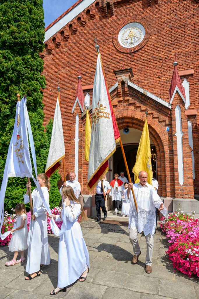 Procesja religijna przed ceglanym kościołem z kolorowymi sztandarami. Jasne stroje uczestników, w tle zielone drzewo. Kwiaty po bokach.