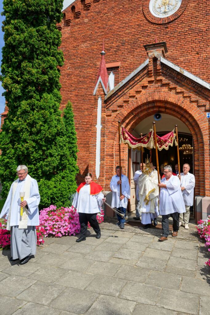 Procesja religijna przed ceglanym kościołem. Uczestnicy ubrani są w białe szaty, trzymają kadzidło i baldachim. Obok rośnie zielony krzew i kwitną różowe kwiaty.