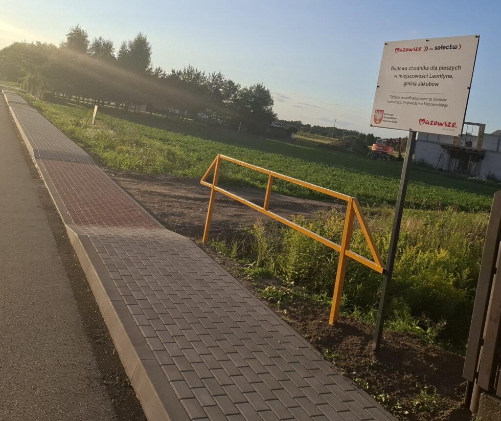 Ścieżka z szarych płytek prowadzi przez zielone pola. Na prawo stoi tablica informacyjna. W tle widać drzewa i budynek.