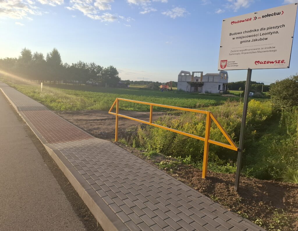 Chodnik z czerwono-szarej kostki prowadzi obok zielonego pola. Po prawej stronie żółta barierka i biały znak z czerwonym napisem. W tle jasne niebo i budynek w budowie.