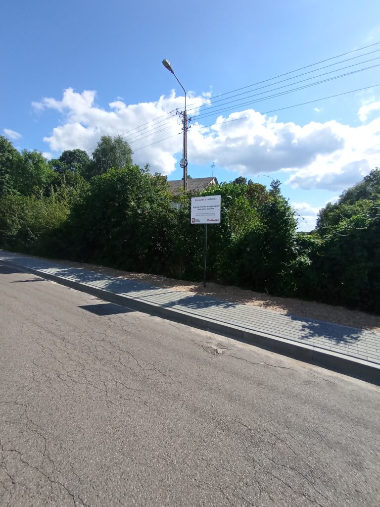 Ulica z chodnikiem obok zielonych krzewów. Jasne niebo z chmurami nad ulicą, lampa i tablica informacyjna. Letnia atmosfera.