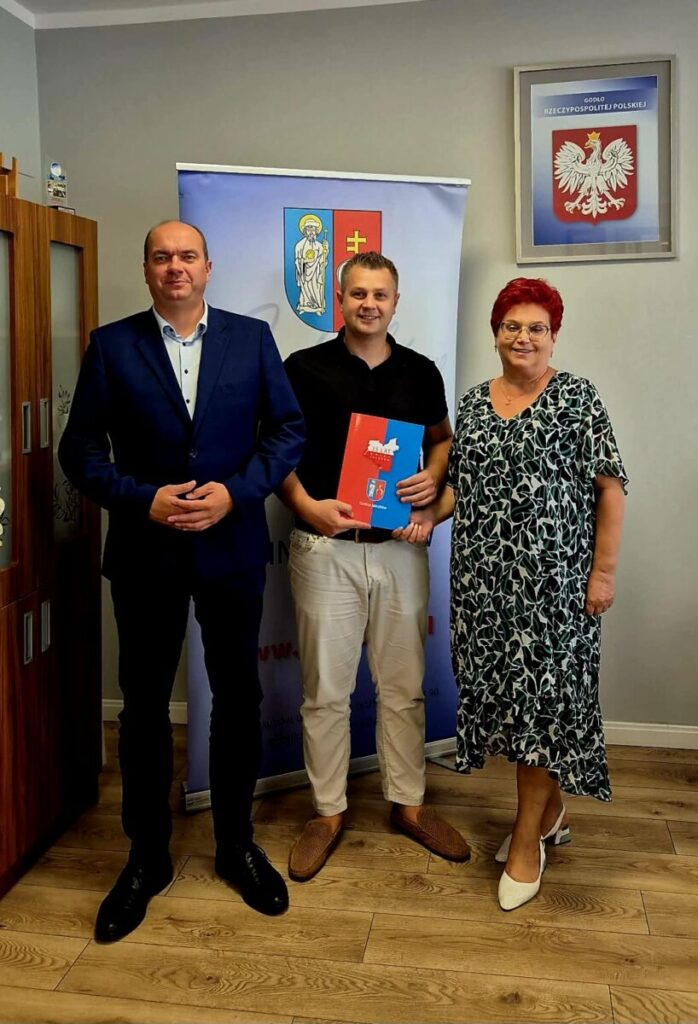 Trzy osoby stoją na tle szarej ściany z godłem Polski. Mężczyzna w środku trzyma kolorową teczkę. Podłoga jest drewniana.