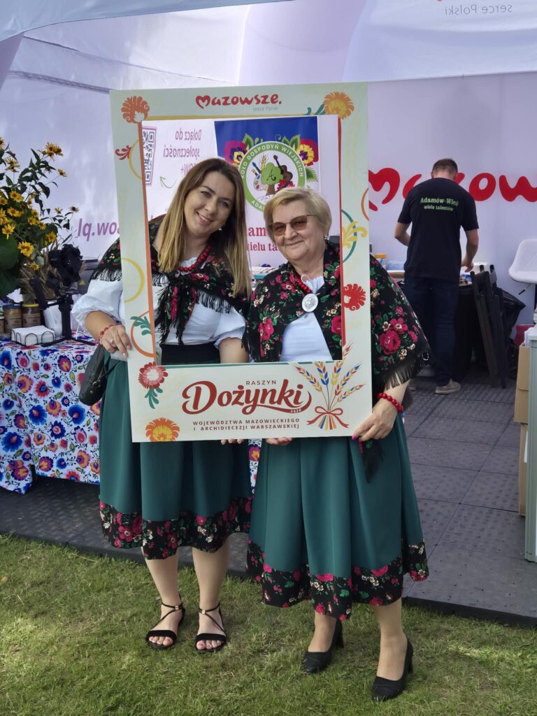 Dwie kobiety w regionalnych strojach ludowych pozują do zdjęcia w ramce. W tle namiot z napisem Mazowsze i dekoracje kwiatowe. Na trawie widoczne stoisko z tradycyjnymi rekwizytami.