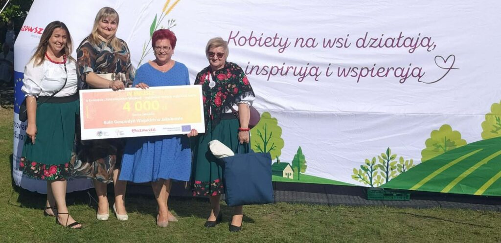 Cztery kobiety pozują z czekiem na trawie. W tle biały baner z napisem o działalności kobiet na wsi. Kobiety ubrane są w kolorowe stroje ludowe.
