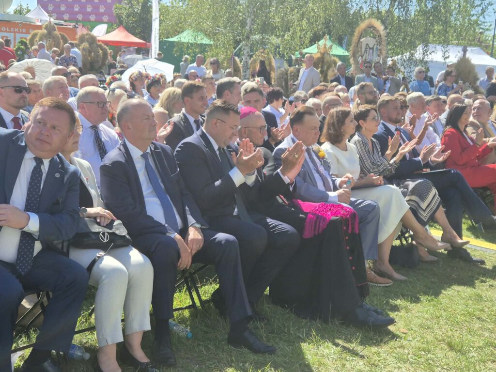 Grupa elegancko ubranych ludzi siedzi na zewnątrz i bije brawo. Tło wypełniają drzewa i kolorowe namioty w słońcu. Panuje atmosfera wydarzenia plenerowego.