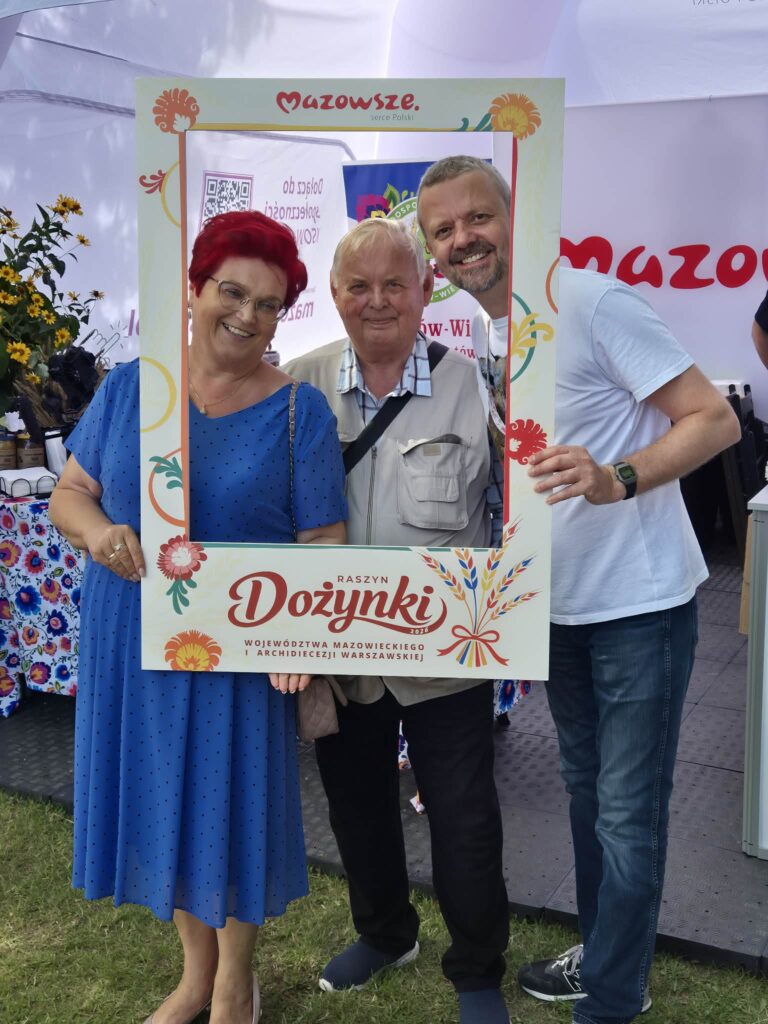 Trzy osoby pozują z ramką Dożynki. Kobieta w niebieskiej sukni, starszy mężczyzna i mężczyzna w białej koszulce. W tle namiot z napisem Mazowsze.