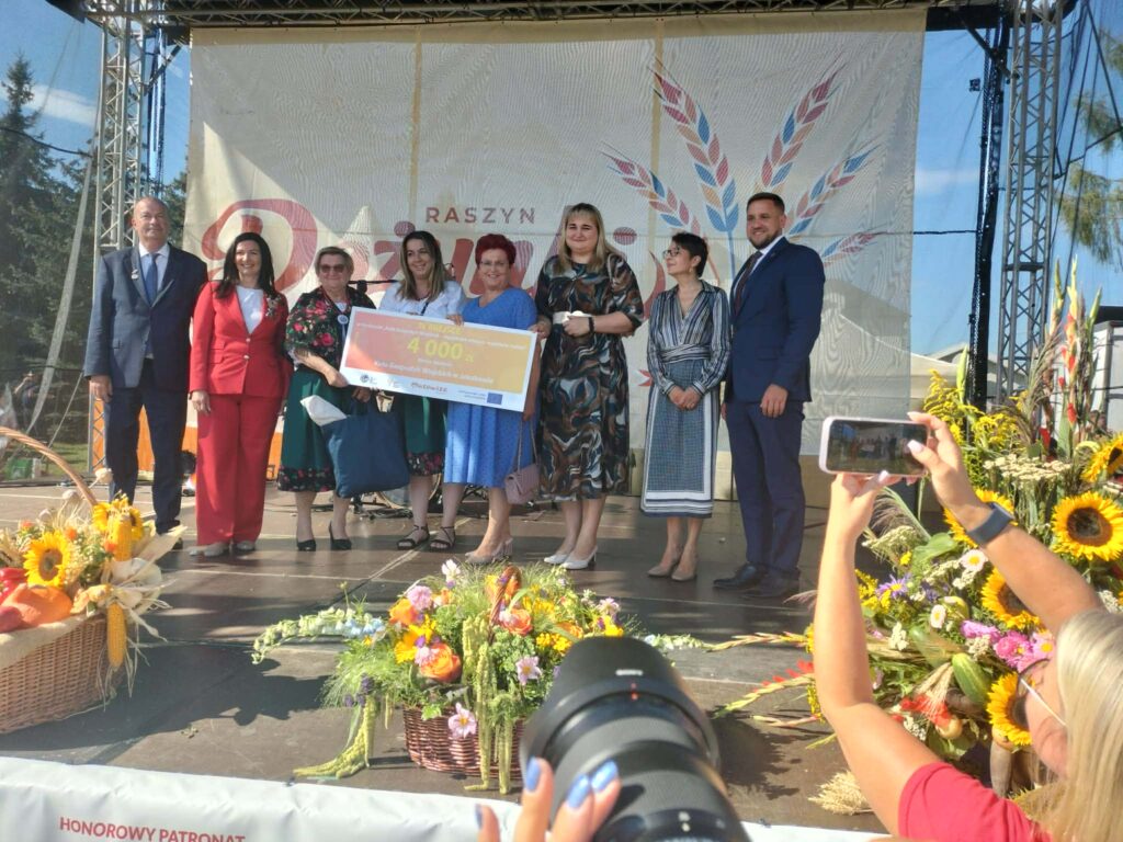 Grupa ludzi na scenie trzyma czek, w tle biały baner z wzorami. Otoczenie ozdobione kwiatami i koszami z dyniami. Dzień jest słoneczny.