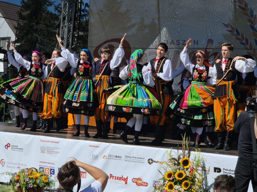 Grupa tancerzy w kolorowych, ludowych strojach tańczy na scenie na tle drzew. Dominują mocne barwy zieleni, pomarańczy i czerni. Przed sceną stoją osoby z aparatami fotograficznymi.
