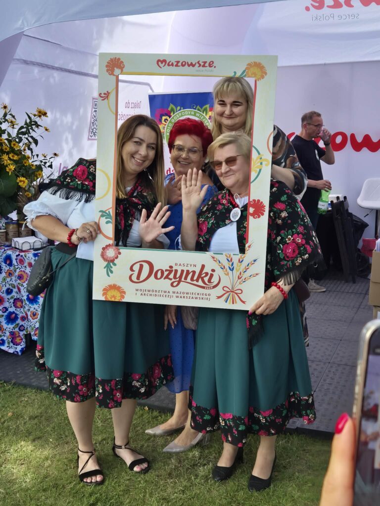 Na zdjęciu cztery osoby w strojach ludowych pozują z ramką do zdjęć na Dożynki w Raszynie. Tło zdobią kwiaty i napis Mazowsze. Dominuje kolor zielony, czerń i czerwone akcenty.