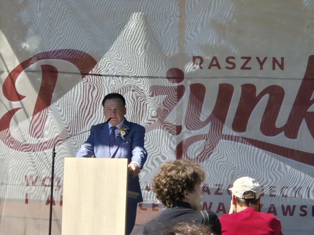 Mężczyzna w niebieskim garniturze przemawia przy mównicy. Tło to baner z napisem RASZYN Dożynki. Obok stoją dwie osoby.