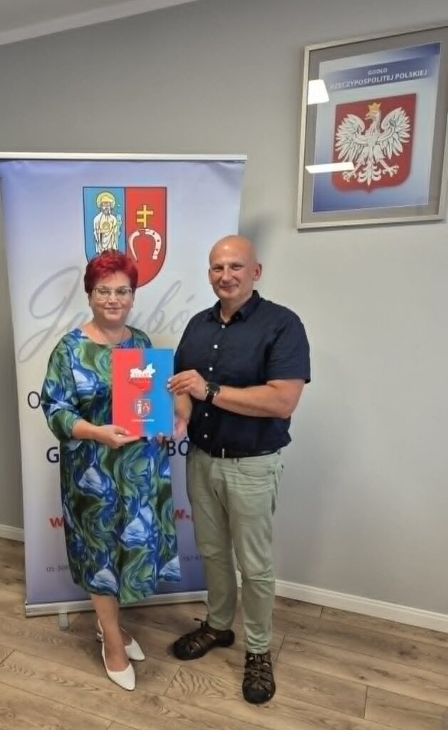 Dwie osoby pozują z kolorową teczką na tle roll-upu z herbem, w jasnym pomieszczeniu z drewnianą podłogą i godłem Polski.
