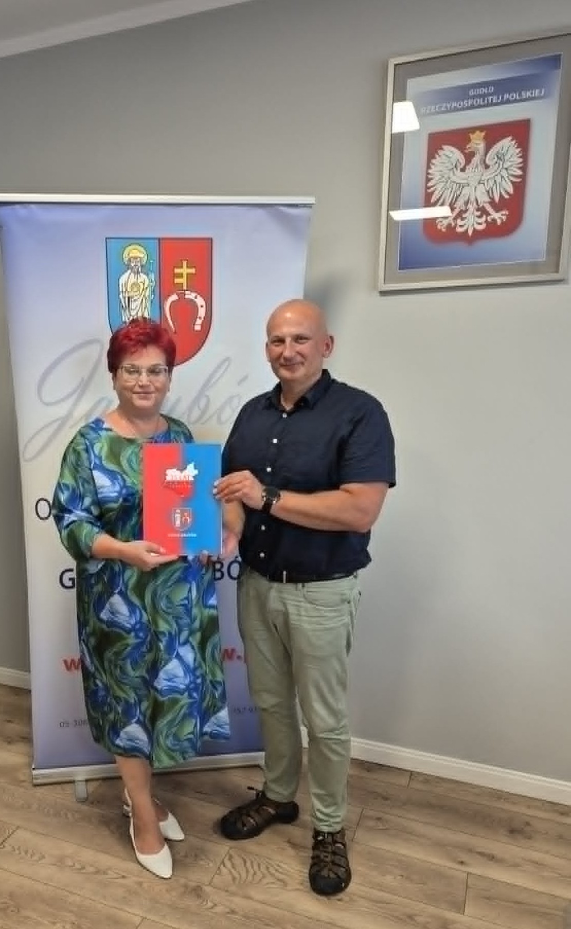 Dwie osoby pozują z kolorową teczką na tle roll-upu z herbem, w jasnym pomieszczeniu z drewnianą podłogą i godłem Polski.