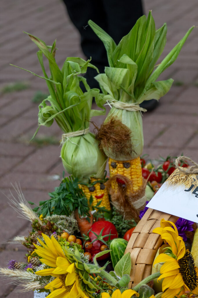 Kosz pełen kolorowych warzyw i kwiatów, ozdobiony kukurydzianymi lalkami. Dominują jaskrawe żółcie i zielenie. Tło to brązowa kostka brukowa.