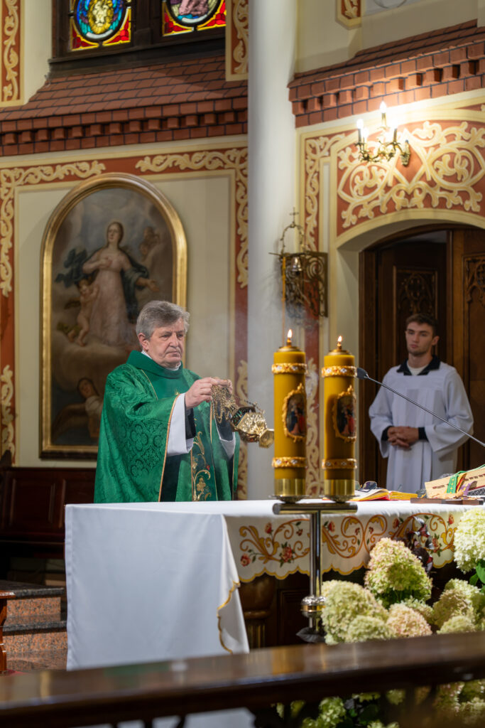 Kapłan w zielonym ornacie używa kadzielnicy w bogato zdobionym kościele. W tle obraz i witraże. Ołtarz ozdobiony kwiatami.