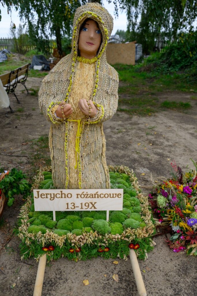 Figura z owsa i słomy przedstawia postać z założonym kapturem. Ma niebieskie oczy, stoi na tle zieleni. Tablica z napisem: Jerycho różańcowe 13-19X.