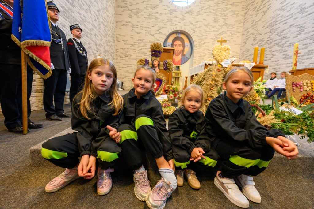Cztery dziewczynki w czarnych strojach z zielonymi wstawkami siedzą przed ołtarzem. W tle dekoracje dożynkowe i obraz. Za nimi strażacy w mundurach.