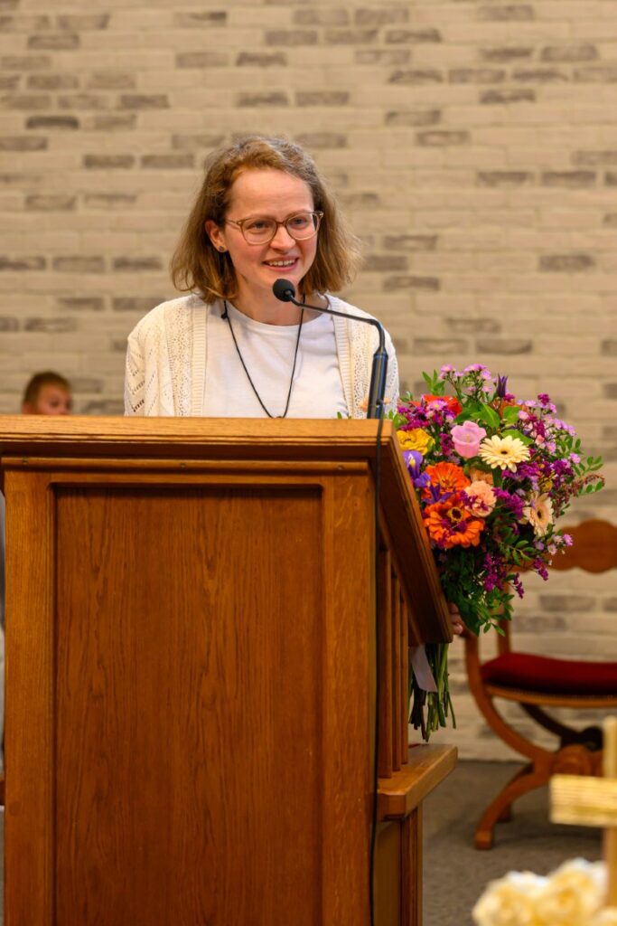 Osoba przy mównicy trzyma bukiet kolorowych kwiatów. Tło to ceglana ściana w jasnych odcieniach. Ubrana w biały strój.