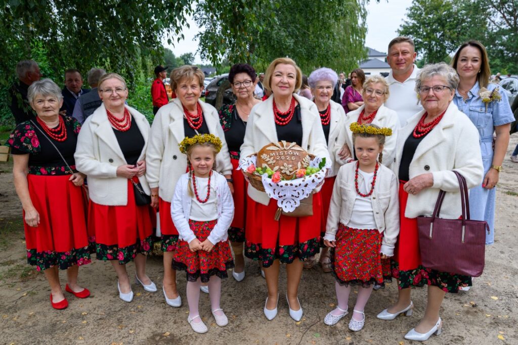 Grupa osób w tradycyjnych, czerwono-białych strojach ludowych na tle drzew. Kobieta z koszem z napisem Miastków 2026. Pogodny dzień.