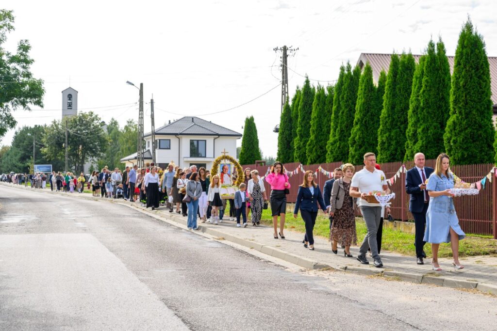Na zdjęciu widać kolorowy pochód ludzi idących ulicą obok zielonych tui i ogrodzenia. W tle biały dom i kościół. Niebo jasne, miejscami zachmurzone.