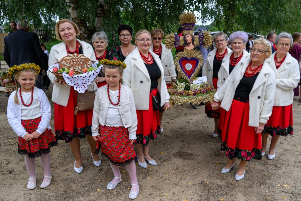 Grupa osób w strojach ludowych, z czerwonymi spódnicami i koralami, stoi na tle zielonych drzew. Kobiety trzymają tradycyjne wieńce dożynkowe. Dzieci mają wianki na głowach.