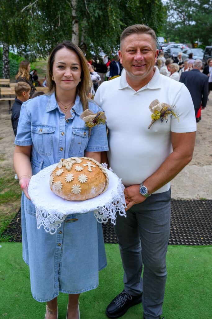 Para w eleganckich strojach trzyma chleb na białej serwecie. W tle trawa i drzewa. Mężczyzna nosi biały sweter, a kobieta niebieską sukienkę.