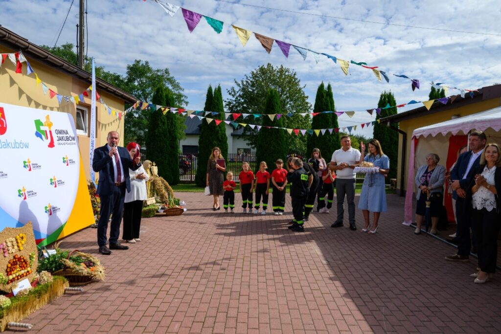 Na zdjęciu widać ludzi na zewnątrz podczas uroczystości z girlandami nad głowami. Stoją na wybrukowanym placu przed budynkiem, dominują kolory zielony i żółty. W tle widać drzewa i niebo z chmurami.