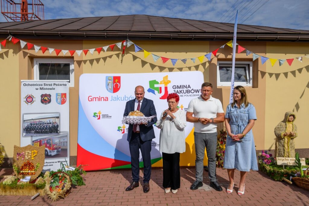 Na tle budynku stoi cztery osoby, jedna trzyma chleb. Baner gminy Jakubów i girlandy w tle. Po lewej stronie jest napis Ochotnicza Straż Pożarna.