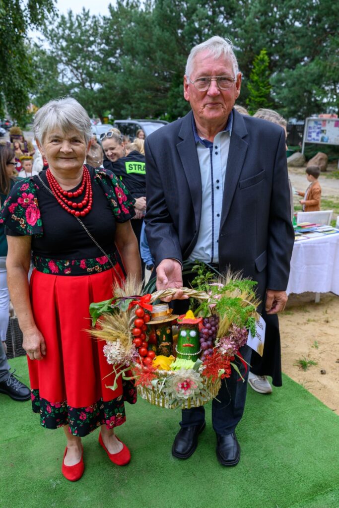 Seniorzy w kolorowych strojach ludowych trzymają kosz z warzywami. W tle drzewa i ludzie. Zielony dywan na ziemi.