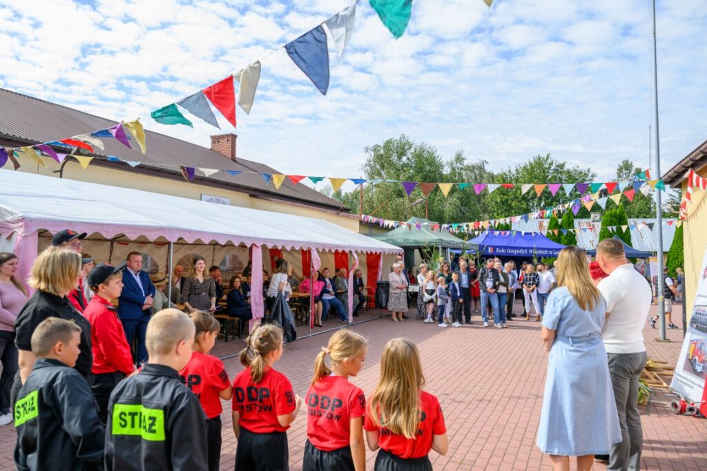 To zdjęcie przedstawia festyn plenerowy z kolorowymi flagami nad głowami ludzi. W tle namiot z siedzącymi osobami, a na pierwszym planie dzieci w czerwonych koszulkach. Dzień jest słoneczny, a niebo częściowo zachmurzone.