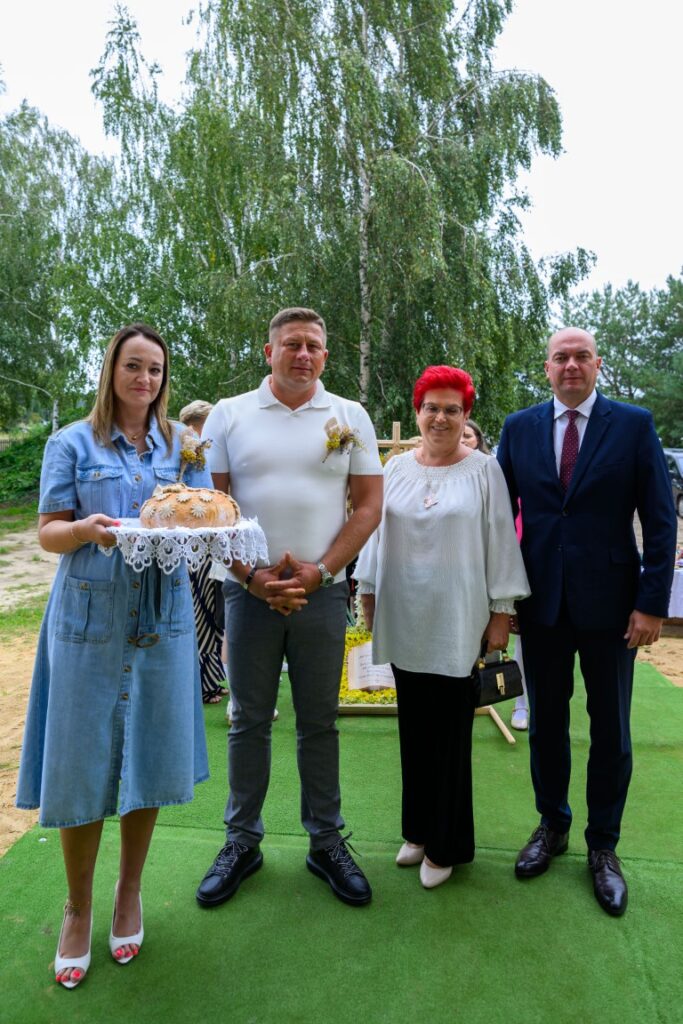 Cztery osoby stoją na trawie, w tle drzewa. Kobieta po lewej trzyma chleb na serwecie. Ubrani elegancko w odcieniach niebieskiego, białego i czerwonego.