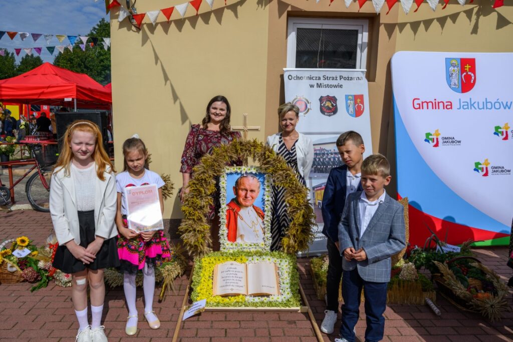 Na zdjęciu widoczna jest grupa dzieci i dorosłych obok portretu papieża otoczonego kwiatami. Tło stanowi kremowa ściana, nad którą wiszą kolorowe flagi. Widać także transparenty z oznakowaniem lokalnym.