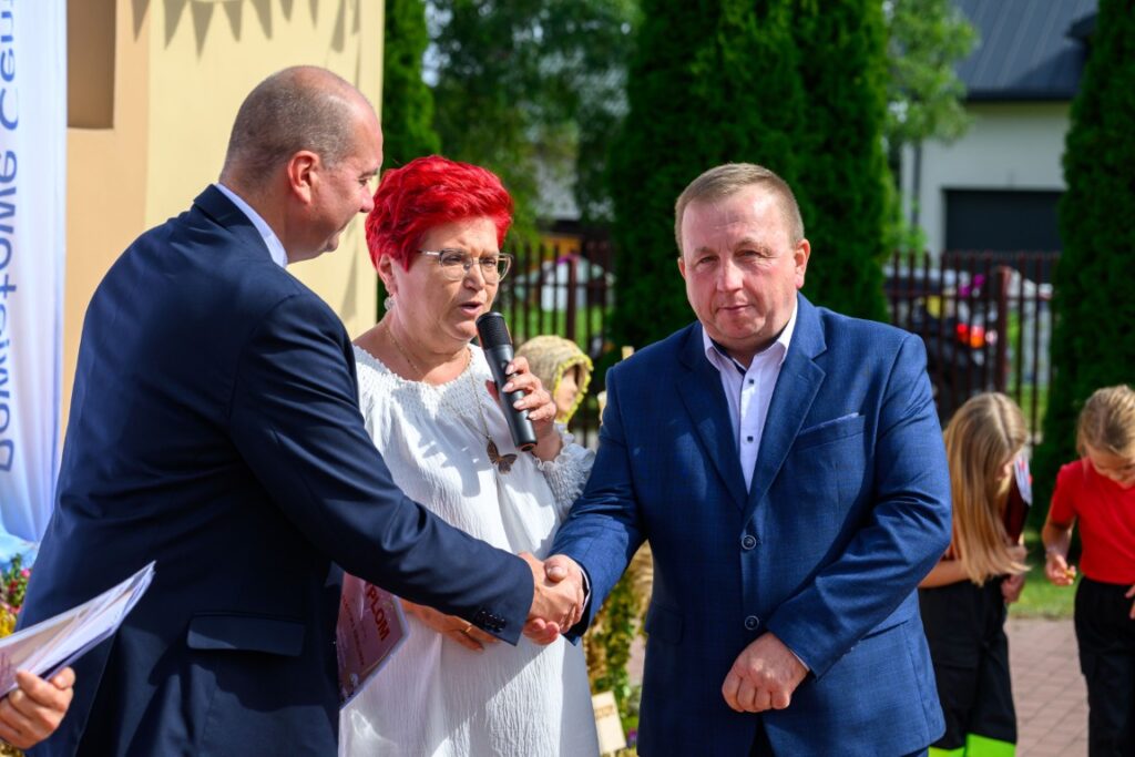Trzy osoby na zewnątrz; mężczyzna w granatowej marynarce, kobieta z czerwonymi włosami, trzyma mikrofon; jasnozielone drzewa w tle.