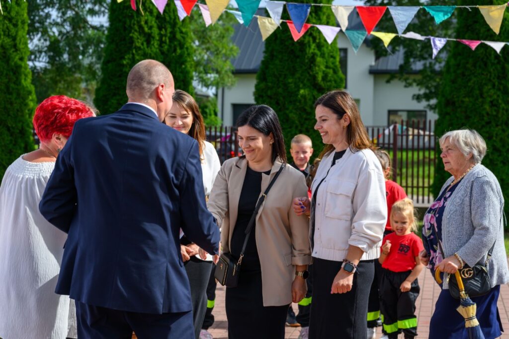 Grupa osób zebrana na wydarzeniu na świeżym powietrzu. Kolorowe chorągiewki wiszą nad nimi. W tle zielone drzewa i budynek.
