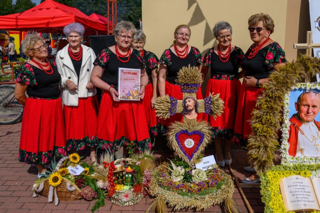 Grupa starszych kobiet w czerwonych spódnicach pozuje z wieńcami dożynkowymi. W tle czerwone namioty. Obok wizerunek papieża.