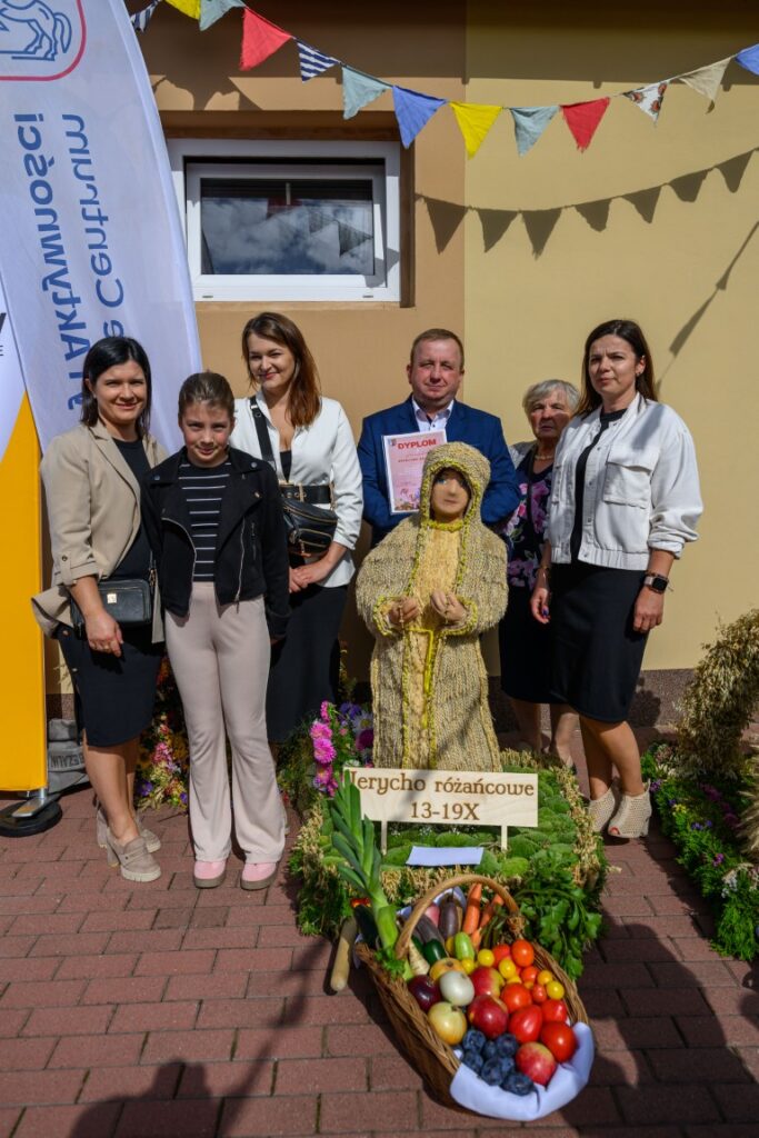 Grupa osób stoi przed budynkiem z beżową fasadą i kolorowymi chorągiewkami. Przed nimi znajduje się koszyk z owocami oraz figura. Jedna osoba trzyma dyplom, obok biały i żółty baner.