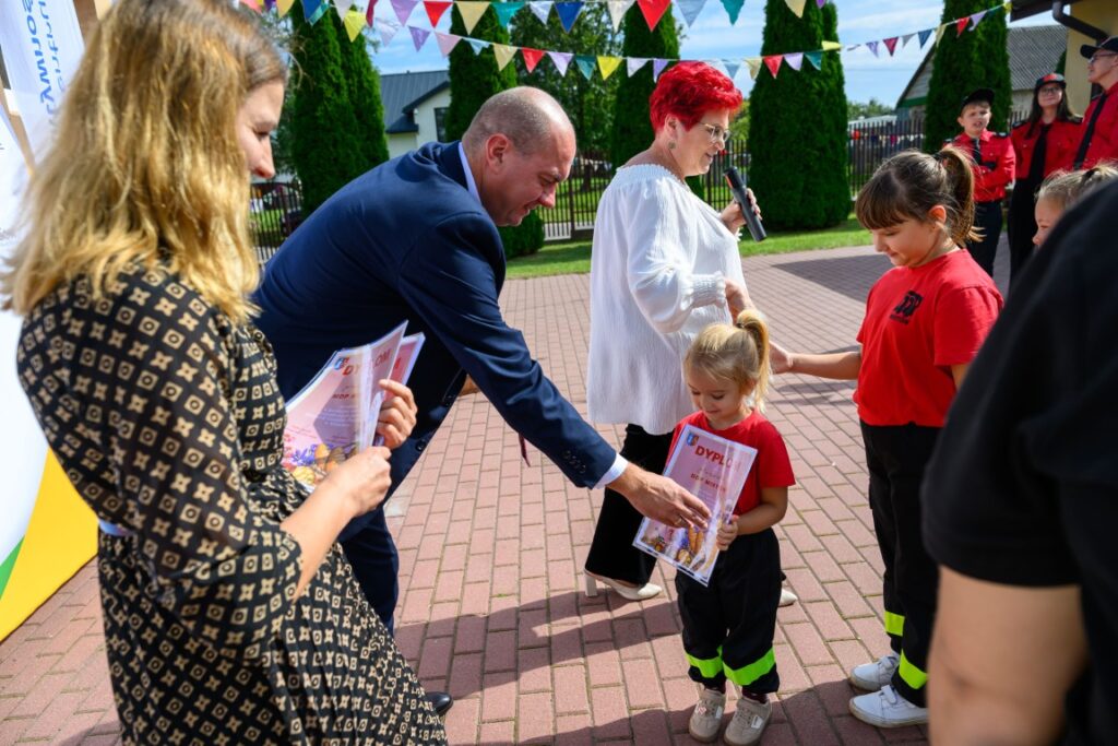 Na zdjęciu mężczyzna wręcza dyplomy dzieciom ubranym w czerwone koszulki. W tle widać kolorowe girlandy i zielone krzewy. Jasne, słoneczne popołudnie, atmosfera jest radosna.