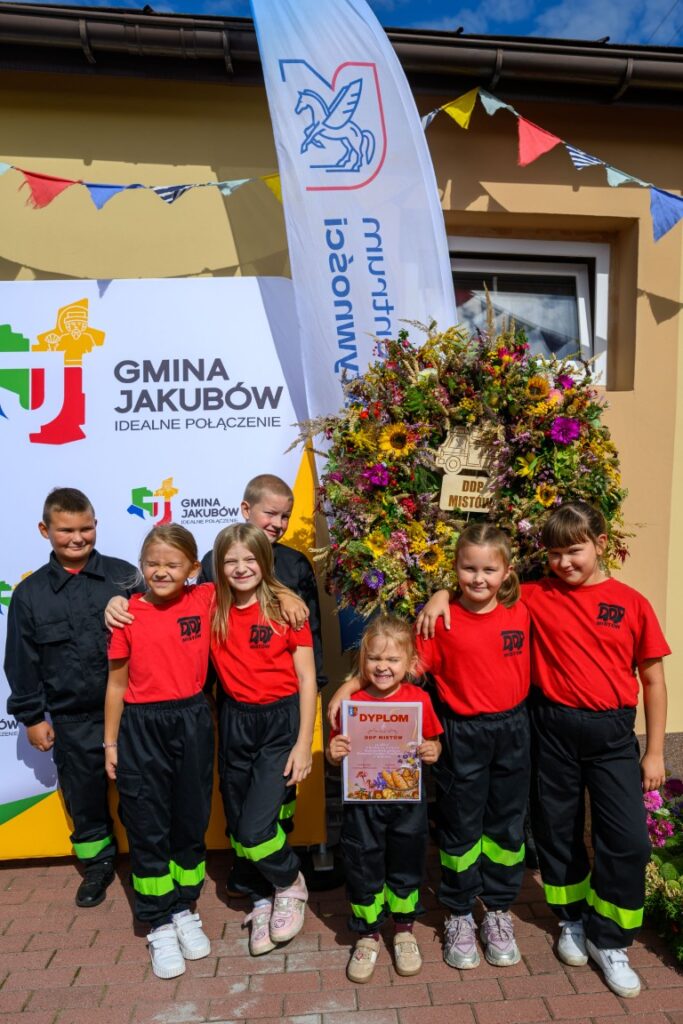 Grupa dzieci w czerwonych koszulkach i czarnych spodniach pozuje z dyplomem. W tle kolorowy wieniec i baner z napisem GMINA JAKUBÓW. Na zewnątrz w pogodny dzień.