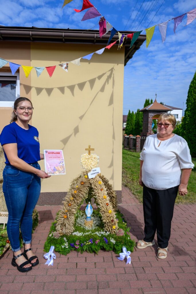 Na zdjęciu dwie kobiety stoją przed ozdobną dekoracją zbożową z figurką Maryi. Tło to budynek w kolorze kremowym z niebieskim niebem. Kolorowe chorągiewki wiszą nad ich głowami.