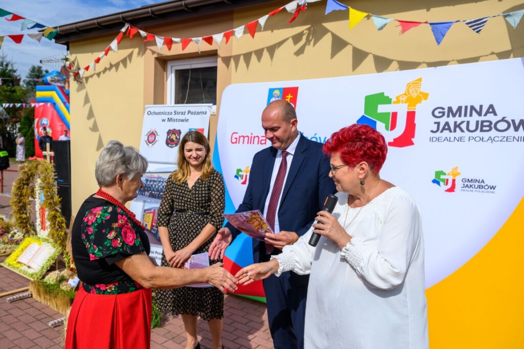 Na zdjęciu widać ceremonię wręczenia dyplomu na tle banerów promujących gminę. Kobieta w czerwonej spódnicy podaje dyplom innemu uczestnikowi. W tle widać kolorowe flagi i żółty budynek.