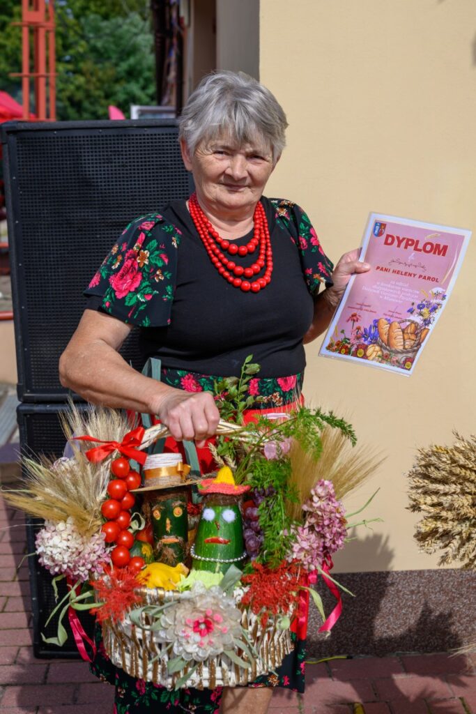 Starsza kobieta w kwiecistej sukience trzyma dyplom i kosz z plonami. Kosz zawiera ozdoby z warzyw i zbożowych elementów. W tle jasna ściana i ciemny głośnik.