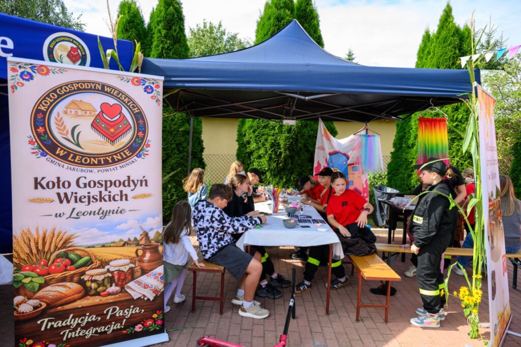 Grupa dzieci tworzy prace plastyczne pod niebieskim namiotem. Obok stoi barwny plakat Koła Gospodyń Wiejskich. Tło to zielone tuje i kolorowe dekoracje.