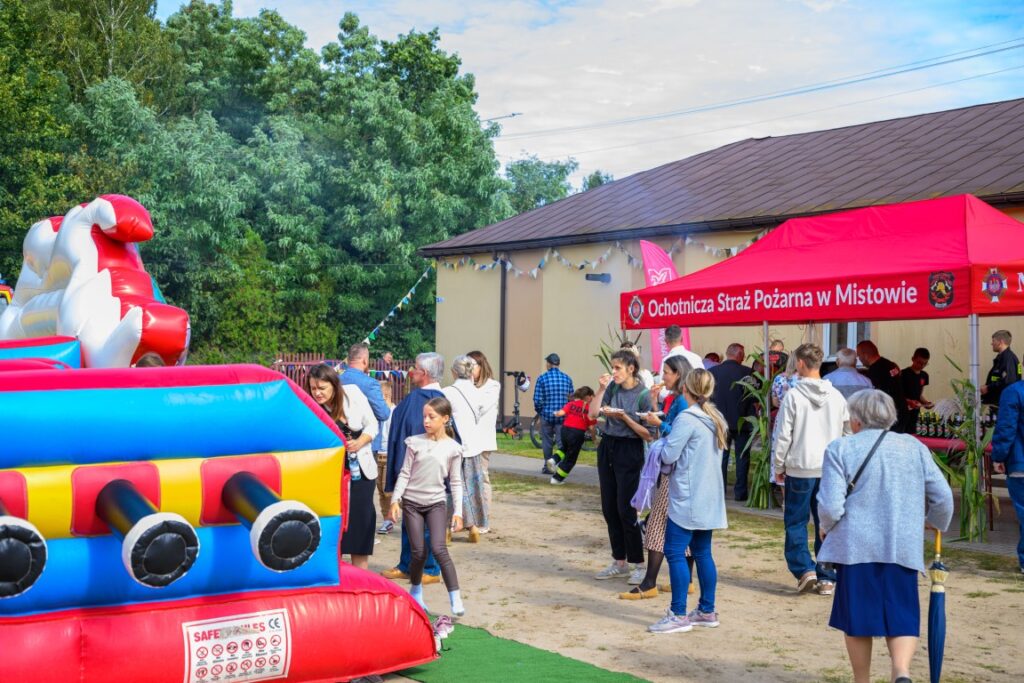 Plac zabaw z dmuchańcem w kolorach czerwonym, żółtym i niebieskim. W tle czerwona altana OSP i grupa osób. Zielone drzewa jako tło.